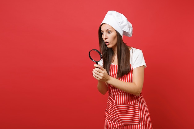 Femme au foyer femme chef cuisinier ou boulanger en tablier rayé t-shirt blanc toque chapeau de chef isolé sur fond de mur rouge. Femme de ménage tenir et regarder à travers une loupe. Maquette concept d'espace de copie