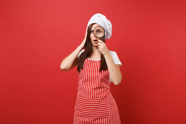 Femme au foyer femme chef cuisinier ou boulanger en tablier rayé t-shirt blanc toque chapeau de chef isolé sur fond de mur rouge. Femme de ménage tenir et regarder à travers une loupe. Maquette concept d'espace de copie