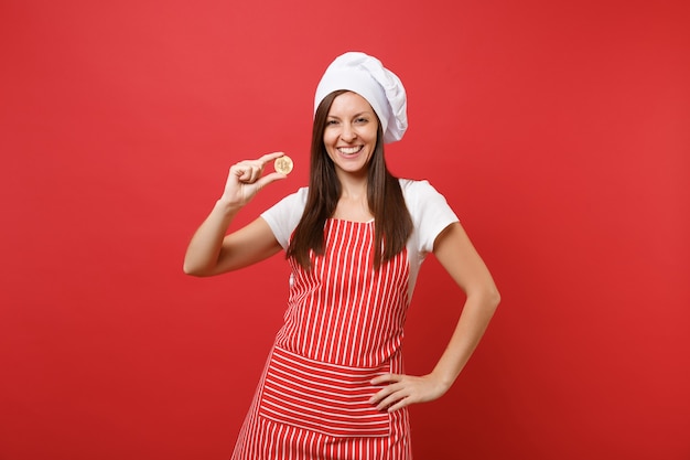 Femme au foyer femme chef cuisinier ou boulanger en tablier rayé t-shirt blanc toque chapeau de chef isolé sur fond de mur rouge. Femme souriante tenant bitcoin, monnaie future bit coin. Maquette du concept d'espace de copie.