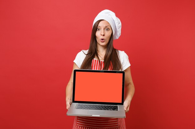 Femme au foyer femme chef cuisinier ou boulanger en tablier rayé t-shirt blanc toque chapeau de chef isolé sur fond de mur rouge. Femme tenir un écran vide pour ordinateur portable pour le contenu promotionnel Maquette concept d'espace de copie