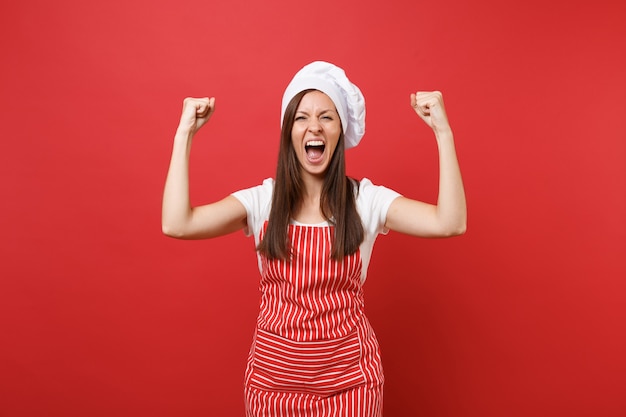 Femme au foyer femme chef cuisinier ou boulanger en tablier rayé t-shirt blanc toque chapeau de chef isolé sur fond de mur rouge. Heureuse femme expressive faisant le geste du gagnant, disant oui. Maquette du concept d'espace de copie.