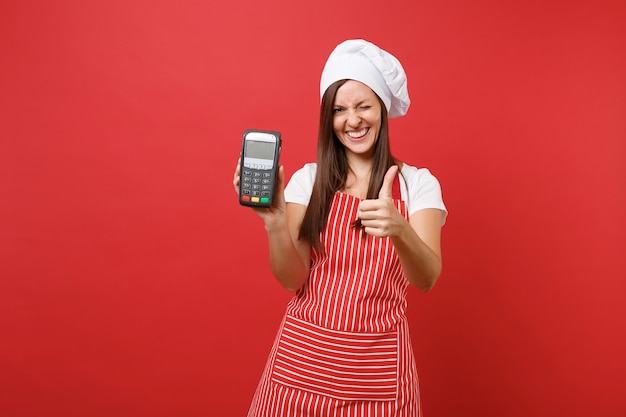 Femme au foyer femme chef cuisinier ou boulanger en tablier rayé t-shirt toque chapeau de chef isolé sur fond de mur rouge. Femme tenir en main un terminal de paiement bancaire sans fil nfc. Maquette du concept d'espace de copie.