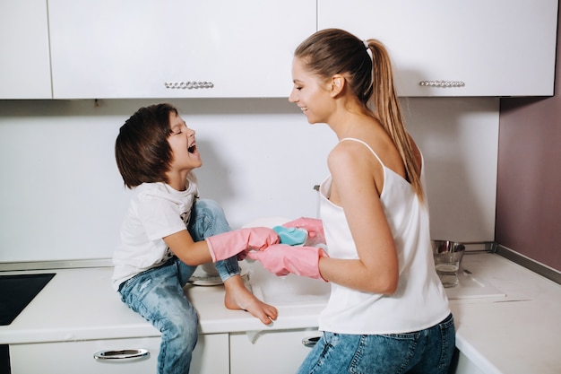 Femme au foyer maman en gants roses lave la vaisselle avec son fils à la main dans l'évier avec un détergent. Une fille en blanc et un enfant avec un plâtre nettoie la maison et lave la vaisselle avec des gants roses faits maison.