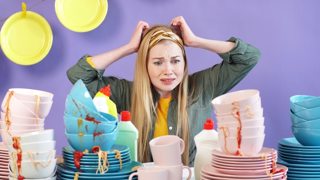 La femme au foyer paniquée tient sa tête dans ses mains et regarde la table avec une montagne d'assiettes et de plats sales non lavés