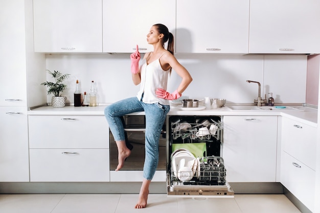 Une femme au foyer portant des gants roses après avoir nettoyé la maison est assise fatiguée dans la cuisine. Dans la cuisine blanche, la fille a lavé la vaisselle et se repose. Beaucoup de vaisselle lavée