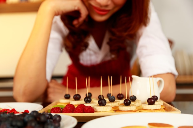 La femme au foyer prépare et décore de délicieux petits fruits mélangés sains, banane, fraise, myrtille et pile d'apéritif aux crêpes aux cerises décorée d'un bâton.