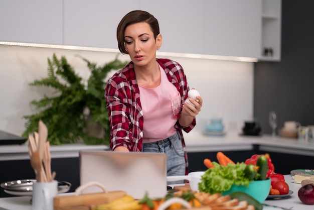 Femme au foyer à la recherche de recettes en ligne à l'aide d'un ordinateur portable pendant la cuisson ou la cuisson des œufs