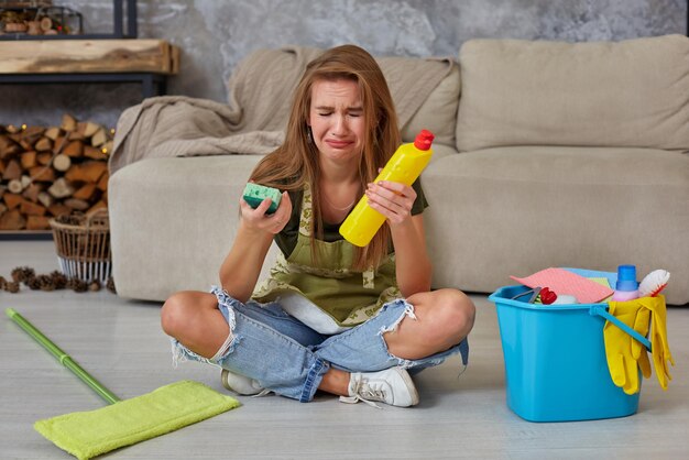 Une femme au foyer se sent fatiguée assise par terre dans le salon après avoir nettoyé à la maison avec un seau bleu rempli de produits chimiques et d'installations pour ranger