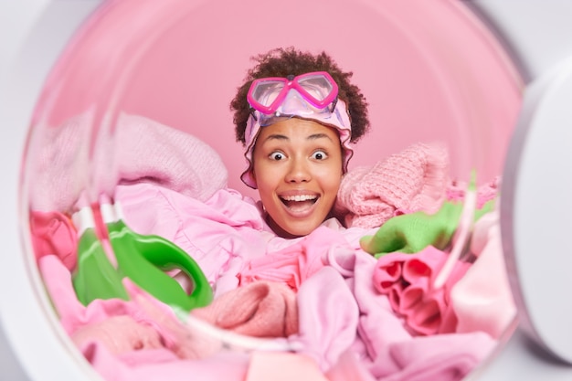 Photo une femme au foyer surprise et heureuse charge une machine à laver automatique avec du linge regarde la caméra ouvre la bouche d'un grand intérêt porte des lunettes de plongée en apnée sur le front enfoui dans un détergent pour vêtements sales près