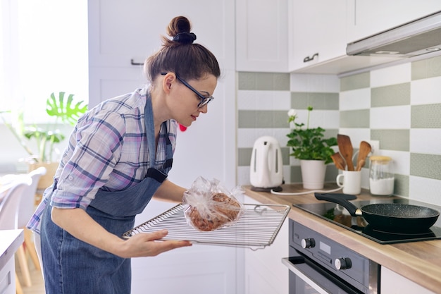 Femme au foyer en tablier préparant du porc dans une manche de cuisson à la maison, met un plateau de viande au four