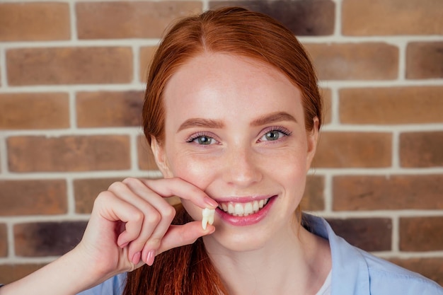 Femme au gingembre rousse avec un sourire blanc comme neige tenant une dent de sagesse blanche après l'ablation chirurgicale d'une dent