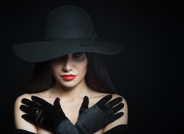 Femme au grand chapeau noir avec les bras sur fond sombre de portrait en studio