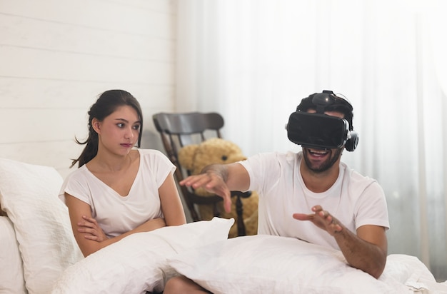 Femme au lit, son mari, homme aux cheveux noirs courts et à la barbe de 3 jours à l'aide de sa tête VR, jeu vidéo avec les lunettes 3D, s'amusant.