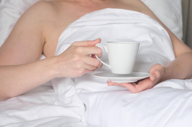 Femme au lit tient une tasse blanche sur une soucoupe dans ses mains