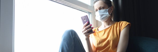 Femme au masque médical de protection assise sur le rebord de la fenêtre avec un téléphone portable dans les mains