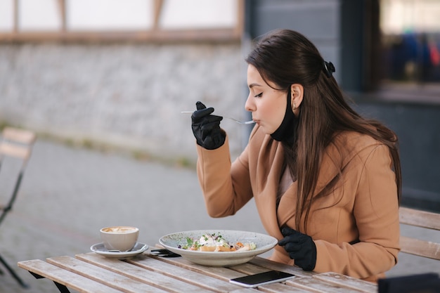 Femme au masque noir assis dans un café et préparé pour manger une salade végétalienne. Concept de café de quarantaine. Covid19.