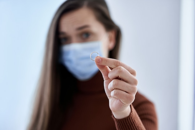 Femme au masque de protection détient l'anneau. Rompre la relation après avoir vécu ensemble et rester à la maison avec son mari pendant la quarantaine et l'isolement en raison de l'épidémie de coronavirus. Concept de divorce