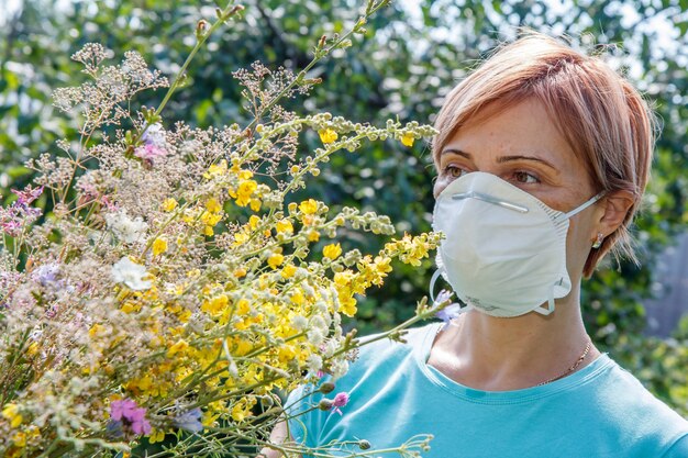 Femme au masque de protection tenant un bouquet de fleurs sauvages et essayant de lutter contre les allergies au pollen. Fond naturel. Notion d'allergie.