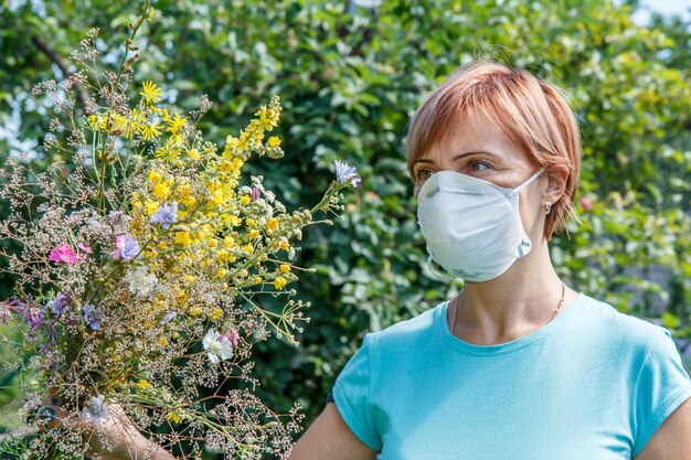 Femme au masque de protection tenant un bouquet de fleurs sauvages et essayant de lutter contre les allergies au pollen. Notion d'allergie.