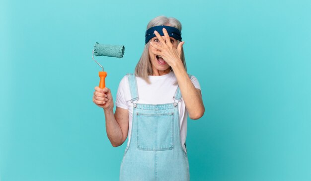 Femme aux cheveux blancs d'âge moyen ayant l'air choquée, effrayée ou terrifiée, couvrant le visage avec la main avec un rouleau peignant un mur