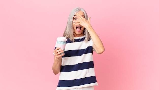 Femme aux cheveux blancs d'âge moyen ayant l'air choquée, effrayée ou terrifiée, couvrant le visage avec la main et tenant un récipient de café à emporter