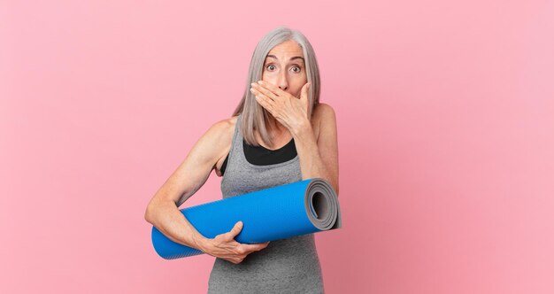 Femme aux cheveux blancs d'âge moyen couvrant la bouche avec les mains avec un choc et tenant un tapis de yoga. concept de remise en forme