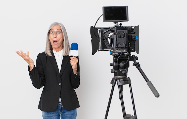 femme aux cheveux blancs d'âge moyen étonnée, choquée et étonnée d'une surprise incroyable et tenant un microphone. concept de présentateur de télévision