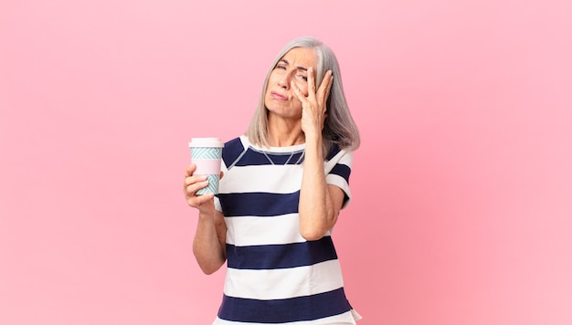 Femme aux cheveux blancs d'âge moyen se sentant ennuyée, frustrée et somnolente après un fastidieux et tenant un récipient de café à emporter