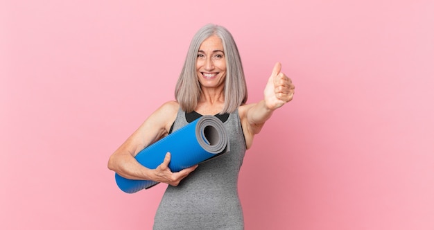 Femme Aux Cheveux Blancs D'âge Moyen Se Sentant Fière, Souriante Positivement Avec Les Pouces Vers Le Haut Et Tenant Un Tapis De Yoga