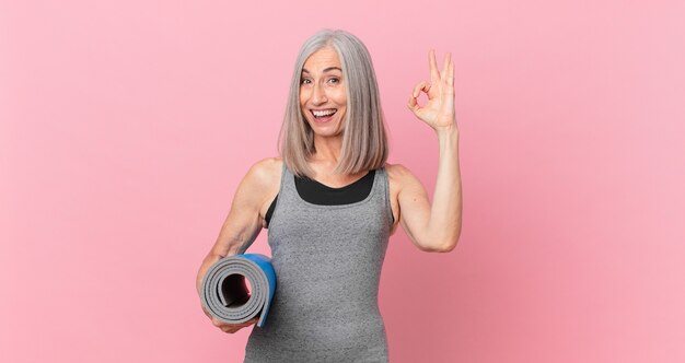 Femme aux cheveux blancs d'âge moyen se sentant heureuse, montrant son approbation avec un geste correct et tenant un tapis de yoga. concept de remise en forme