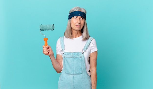 Femme aux cheveux blancs d'âge moyen se sentant triste, contrariée ou en colère et regardant sur le côté avec un rouleau peignant un mur