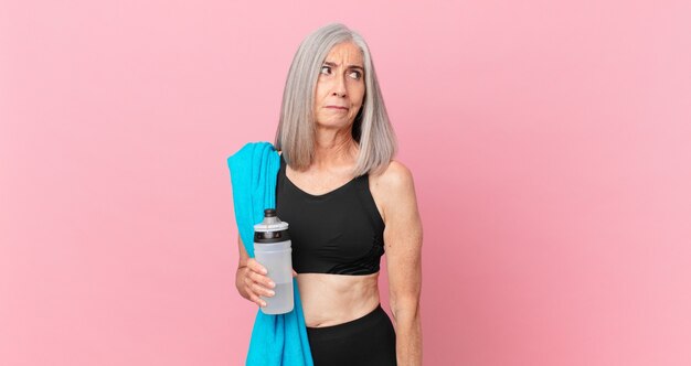 Femme aux cheveux blancs d'âge moyen se sentant triste, contrariée ou en colère et regardant de côté avec une serviette et une bouteille d'eau. concept de remise en forme