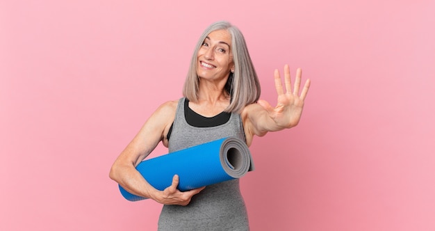 Femme aux cheveux blancs d'âge moyen souriante et semblant amicale, montrant le numéro cinq et tenant un tapis de yoga. concept de remise en forme