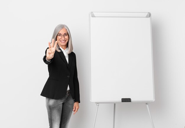 femme aux cheveux blancs d'âge moyen souriante et semblant amicale, montrant le numéro trois et un espace de copie du tableau. concept d'entreprise