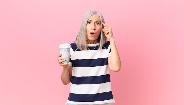 Femme aux cheveux blancs d'âge moyen à la surprise de réaliser une nouvelle idée ou un concept de pensée et tenant un récipient de café à emporter