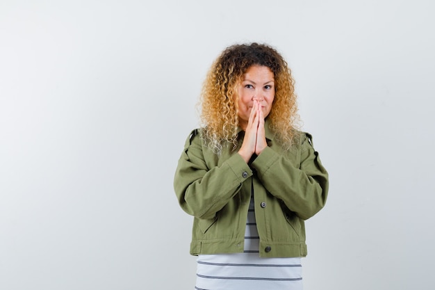 Photo femme aux cheveux blonds bouclés en veste verte en gardant les mains en signe de prière et à la vue de face, pleine d'espoir.