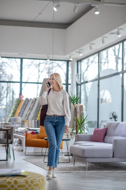 Photo femme aux cheveux blonds parlant sur le smartphone, regardant sur le côté, marchant dans le grand hall, souriant.