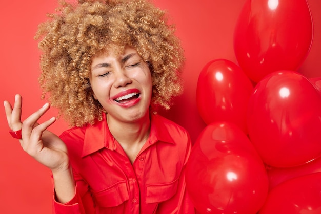 Une femme aux cheveux bouclés bouleversée avec une expression misérable garde la main levée a une fuite de maquillage porte une chemise tient des ballons cardiaques gonflés vêtus d'une chemise isolée sur fond rouge Concept de célébration