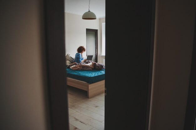 Femme aux cheveux bouclés commençant le matin suivant des cours en ligne sur un ordinateur portable depuis le lit encore en désordre ...