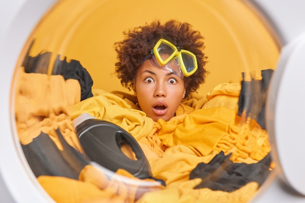 Une femme aux cheveux bouclés effrayée porte un masque de plongée en apnée sur le front noyé dans le linge avec une bouteille de détergent pose contre le mur jaune