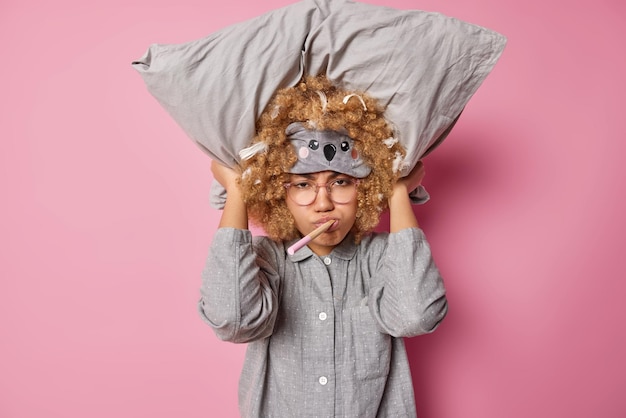 Une femme aux cheveux bouclés insatisfaite garde un oreiller sur la tête porte un masque de sommeil et un pyjama se prépare au sommeil isolé sur fond rose garde une brosse à dents en bois dans la bouche Sommeil repos et heure du coucher