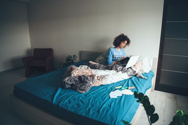 Femme aux cheveux bouclés lisant un livre le matin assis dans son lit avec un ordinateur portable