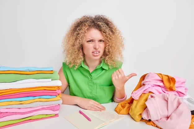 Une femme aux cheveux bouclés mécontente pointe avec le pouce sur le linge déplié se sent fatiguée du travail domestique prend des notes dans le bloc-notes note la température de lavage pour les vêtements faits de matériaux différents.