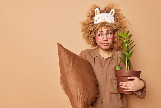 Une femme aux cheveux bouclés mécontente vêtue d'un pyjama confortable tient un pot de plante domestique et un oreiller applique des patchs de beauté sous les yeux isolés sur un espace de copie de fond beige pour votre promotion