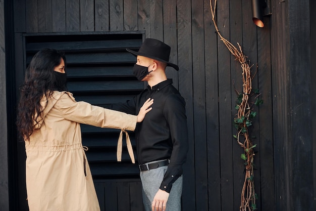 Femme aux cheveux bouclés noirs debout contre l'extérieur du bâtiment en bois noir avec son homme au chapeau