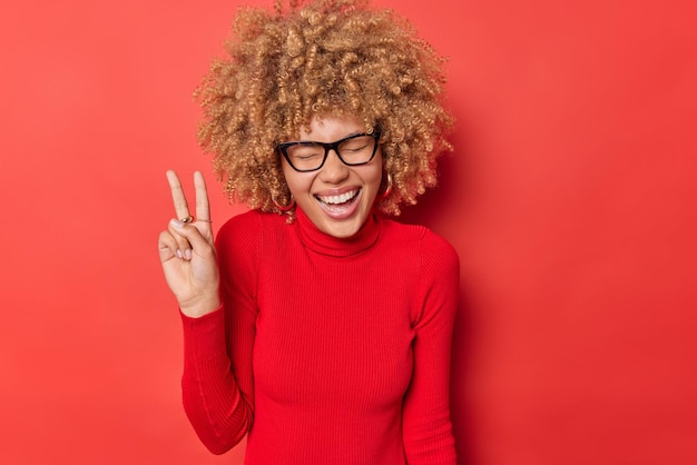 Une femme aux cheveux bouclés ravie rit joyeusement fait un geste de paix aime la vie garde les yeux fermés étant de bonne humeur porte des lunettes et un col roulé isolé sur fond rouge Concept de langage corporel