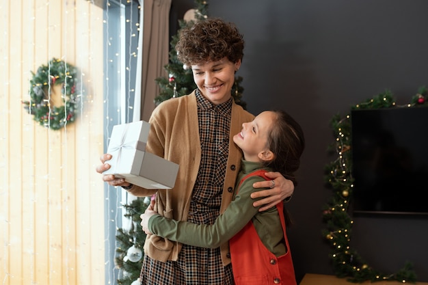 Femme aux cheveux bouclés souriante embrassant sa fille tout en recevant un cadeau de Noël d'elle à la maison