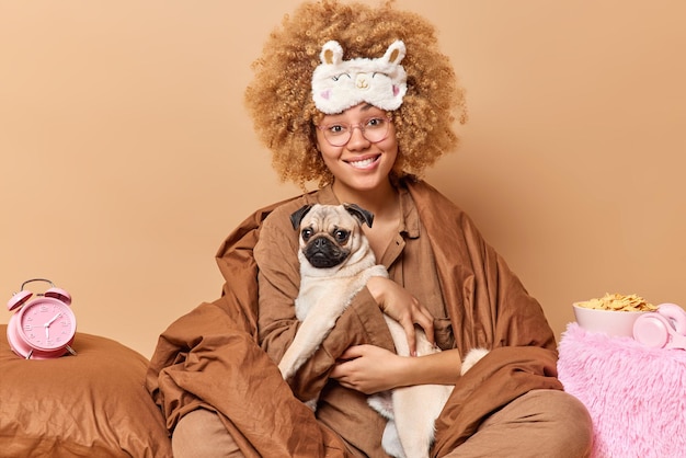 Une femme aux cheveux bouclés souriante et positive enveloppée dans une couverture porte un chien carlin sur les mains reste au lit va dormir profite d'une atmosphère domestique confortable pose sur fond beige Concept de temps en famille