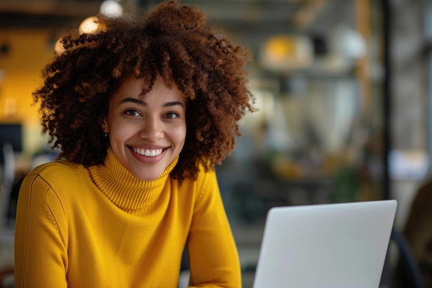 Une femme aux cheveux bouclés sourit et est assise devant un ordinateur portable elle porte un pull jaune et elle est heureuse