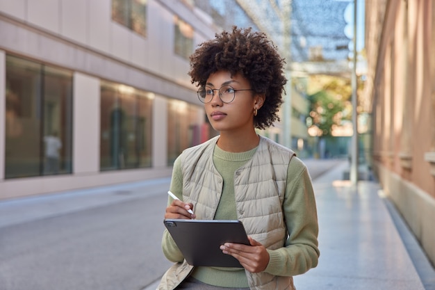 une femme aux cheveux bouclés tient une tablette numérique dessine des croquis avec un stylet regarde ailleurs porte des lunettes rondes un pull décontracté et un gilet crée une illustration sur un appareil numérique